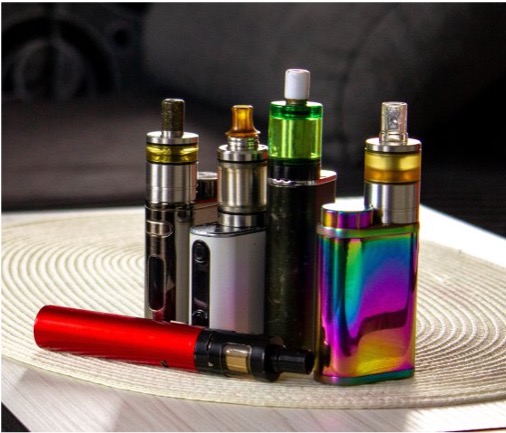 A group of colorful vaping devices in various sizes and designs stand on a woven mat, creating a vibrant and modern display.